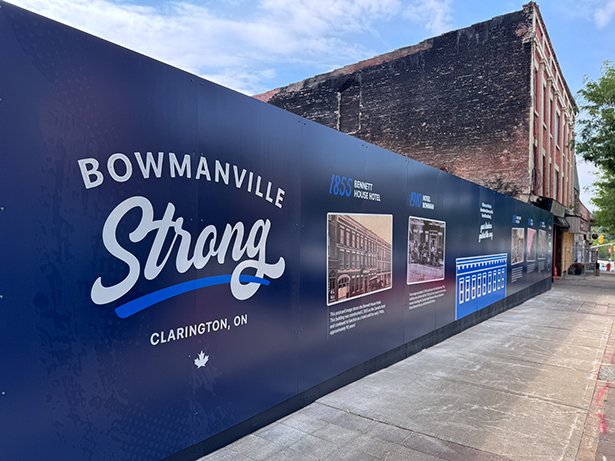 A large blue mural on a sidewalk reads 'Bowmanville Strong, Clarington, ON' with a small maple leaf icon. The mural features historical images and descriptions of buildings from 1885 and 1914, including the Bennett House Hotel and Hotel Centralia. In the background, an old brick building with visible wear is seen.