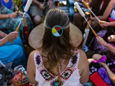 Sacred drums during spiritual singing. A high angle view of a woman wearing native headband and colourful clothes during a singing circle of people around a sacred mother drum outdoors.