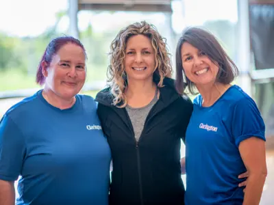 Three Community Services staff standing indoors on the pool deck. Two wear blue 'Clarington' shirts; one wears a black jacket. Green scenery visible through windows.